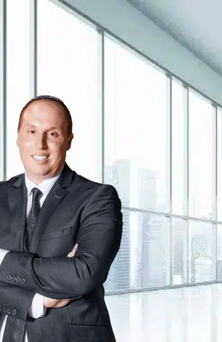 איש עסקים חרדי מחייך לובש חליפה שחורה ועומד במשרד מודרני עם חלונות גדולים.